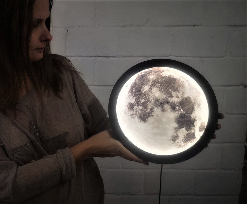 May include: A black and white moon-shaped night light with a realistic moon surface texture. The light is turned on and illuminates the room.