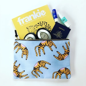 Puede incluir: Una bolsa azul con cremallera con un patrón repetido de tigres naranjas sobre un fondo azul claro. La bolsa está llena de artículos de viaje esenciales, como una revista, un pasaporte, auriculares y artículos de tocador.