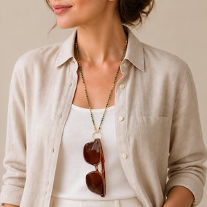 May include: A long necklace featuring small green beads and gold-toned accents, ending in a circular gold-toned ring. A pair of brown-tinted sunglasses with a plastic frame is suspended from the ring, hanging vertically against a white top.