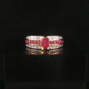 May include: A gold ring featuring a central oval-cut red ruby flanked by two rows of smaller round-cut red rubies. The band is accented with two parallel rows of small white diamonds set along the top and bottom edges of the ruby center.