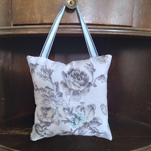 May include: A small white pillow with a floral pattern in shades of gray and blue. The pillow has a lace trim and is hanging from a blue ribbon.