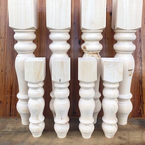 May include: Eight unfinished wooden table legs with a turned design. The legs are various sizes and are arranged in a row against a wood background.