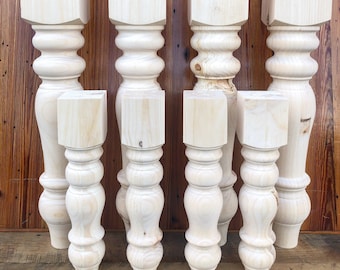 May include: Eight unfinished wooden table legs with a turned design. The legs are various sizes and are arranged in a row against a wood background.