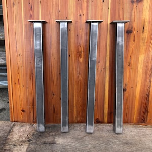 May include: Four silver metal table legs with square tops. The legs are standing upright against a wooden wall.