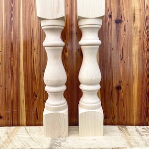 May include: Two unfinished wooden table legs with a turned design. The legs are made of light wood and have a square base.