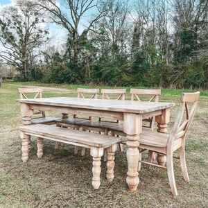 May include: A rustic farmhouse style dining table and bench set made of light wood with a distressed white finish. The table has a rectangular top and six chairs, while the bench has a long, rectangular seat. The set is outdoors on a grassy lawn.