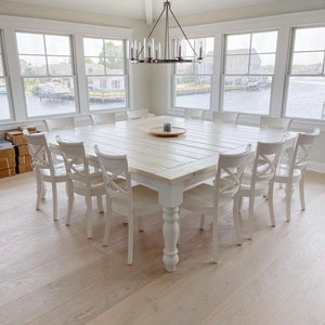 May include: A large, white farmhouse style dining table with a whitewashed wood top and turned legs. The table is surrounded by white chairs with a cross-back design. The table is set with a wooden serving tray and a small plant.
