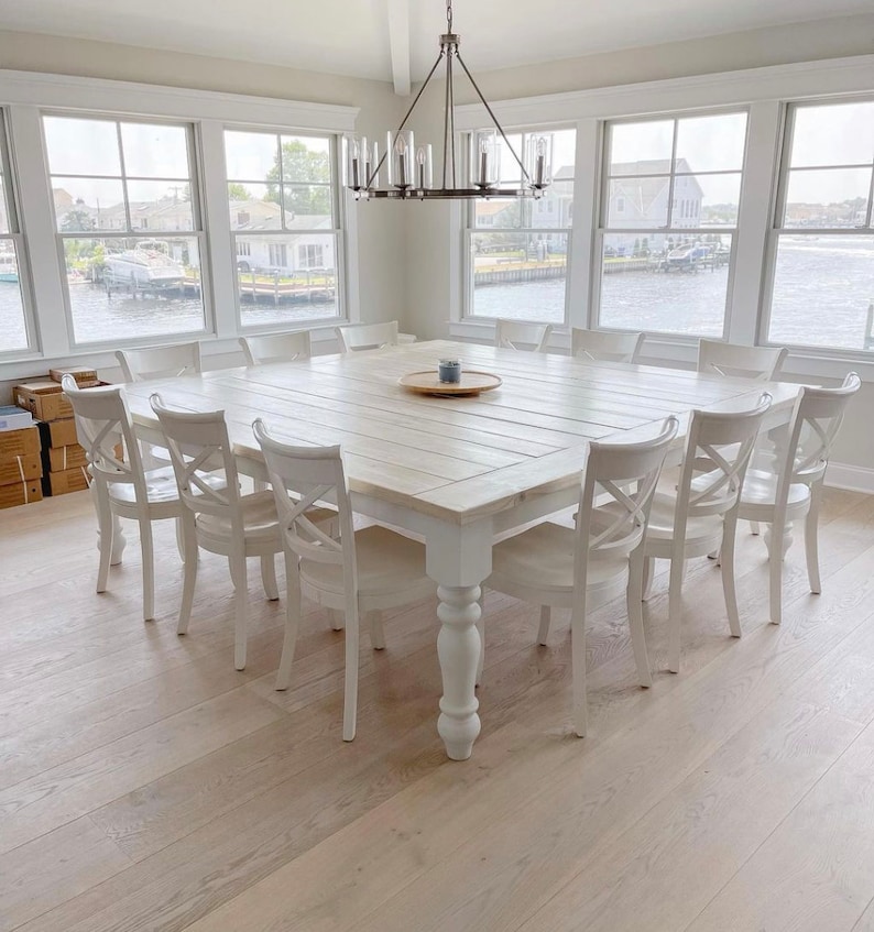 May include: A large, white farmhouse style dining table with a whitewashed wood top and turned legs. The table is surrounded by white chairs with a cross-back design. The table is set with a wooden serving tray and a small plant.