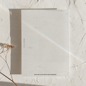 May include: A rectangular sheet of graph paper with a grid pattern, featuring the text "PAPER STANDARDS" and "BIG PLANS. GOOD IDEAS. HIGH STANDARDS." A rainbow prism effect crosses the paper. The paper is on a textured white surface.