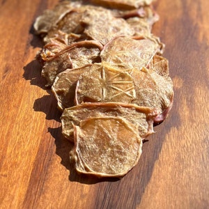 May include: A stack of thin, dried, light brown chicken jerky treats on a wooden surface.