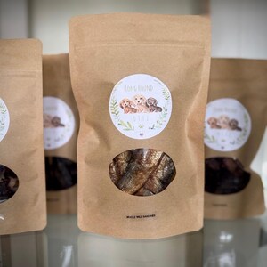 May include: A brown paper bag with a clear window showing a pile of dried fish. The bag has a label with the text "Long Hound Delights" and an illustration of three dachshund puppies. The text "Whole Wild Sardines" is printed below the window.