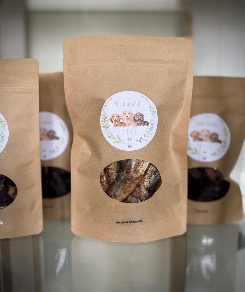 May include: A brown paper bag with a clear window showing a pile of dried fish. The bag has a label with the text "Long Hound Delights" and an illustration of three dachshund puppies. The text "Whole Wild Sardines" is printed below the window.