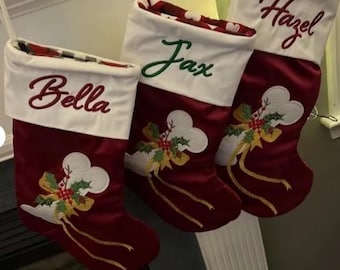 May include: Three maroon Christmas stockings with white cuffs, each featuring embroidered names: Bella, Jax, and Hazel. The stockings display a white bone-shaped applique decorated with green holly leaves, red berries, and gold ribbons.