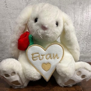 May include: A plush white bunny with long ears and a pink nose. It has a red rose and green leaves attached. A white heart-shaped patch with the name "Evan" embroidered in gold thread and a gold heart is sewn on the bunny.