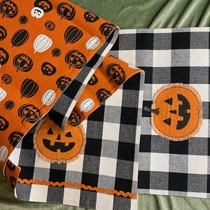 May include: A reversible table runner featuring an orange side with a repeating pattern of black and white pumpkins and a black and white buffalo check side. The buffalo check side includes two orange jack-o'-lantern appliqués and orange trim.