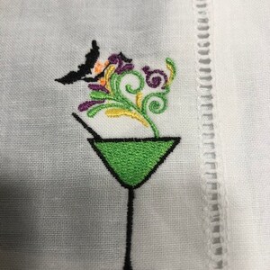May include: White cocktail napkin with an embroidered design. The design features a green martini glass with a black stem, a black bat, and colorful floral accents in purple, orange, and green. The napkin has a decorative stitched border.