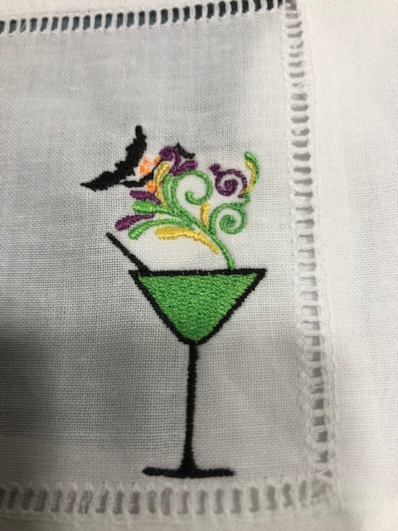 May include: White cocktail napkin with an embroidered design. The design features a green martini glass with a black stem, a black bat, and colorful floral accents in purple, orange, and green. The napkin has a decorative stitched border.