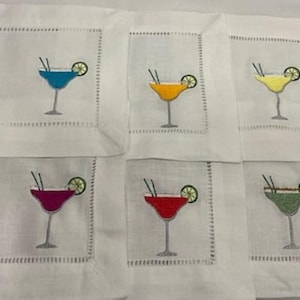 May include: Set of six white linen cocktail napkins, each embroidered with a different colored margarita. The embroidered drinks include blue, yellow, orange, red, green, and purple, each with a lime wedge and straw.