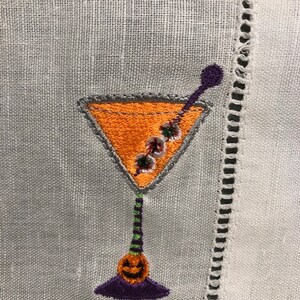 May include: Embroidered cocktail napkin featuring a Halloween-themed martini glass. The glass is orange with a gray outline, a purple stem, and a jack-o'-lantern base. The napkin is white with a decorative stitched border.