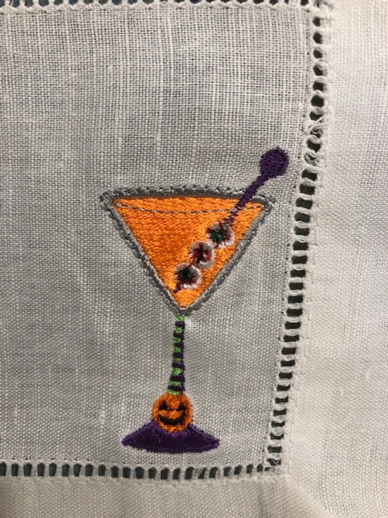 May include: Embroidered cocktail napkin featuring a Halloween-themed martini glass. The glass is orange with a gray outline, a purple stem, and a jack-o'-lantern base. The napkin is white with a decorative stitched border.