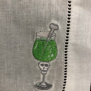 May include: Embroidered cocktail napkin featuring a green drink in a skull-shaped glass. The drink has a white foamy top and a bone-shaped stirrer. The napkin is white with a decorative stitched border.