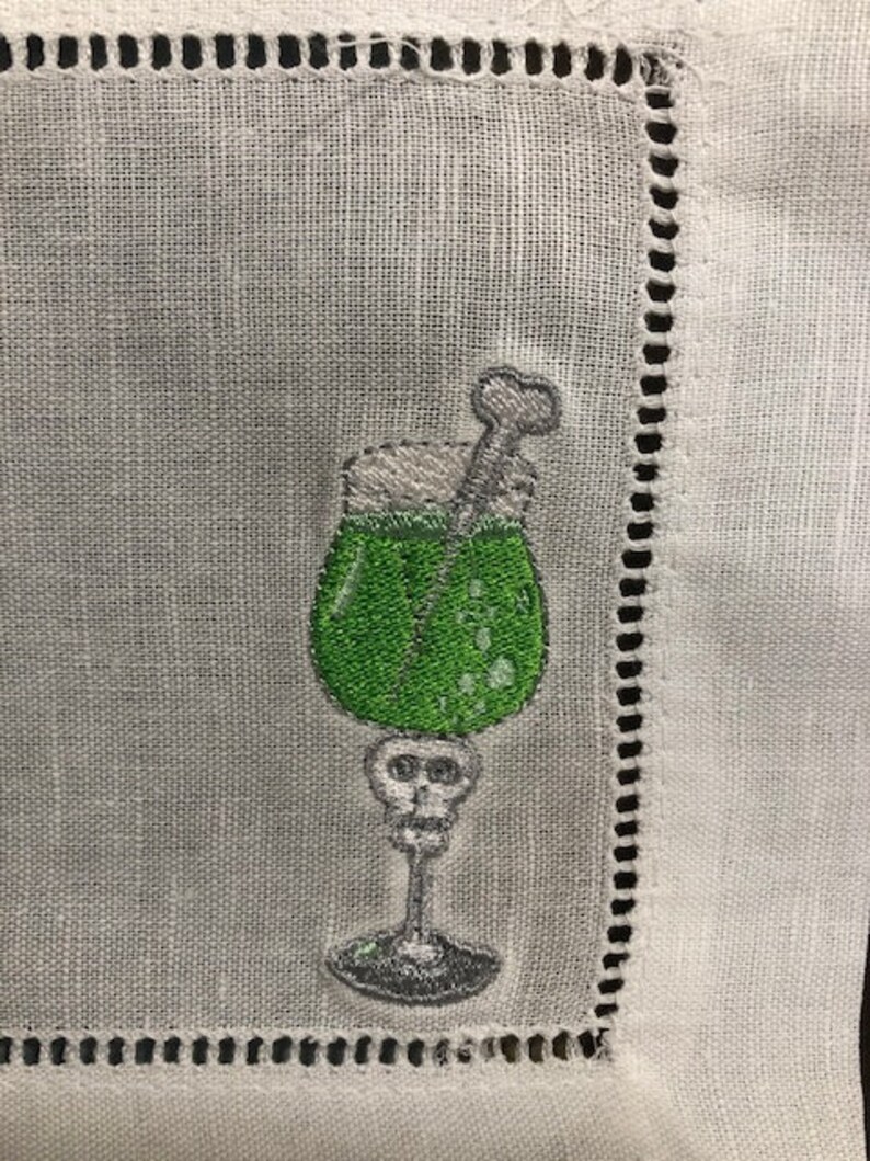 May include: Embroidered cocktail napkin featuring a green drink in a skull-shaped glass. The drink has a white foamy top and a bone-shaped stirrer. The napkin is white with a decorative stitched border.
