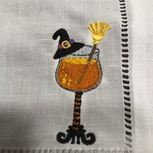 May include: White linen cocktail napkin with an embroidered Halloween design. The design features a cocktail glass filled with an orange liquid, a black witch's hat with a purple band, and a yellow broom. The napkin has a decorative stitched border.