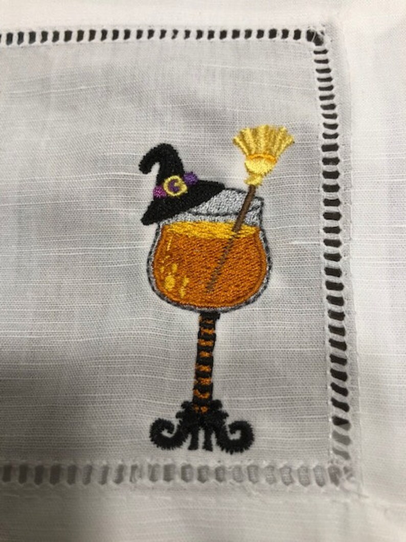 May include: White linen cocktail napkin with an embroidered Halloween design. The design features a cocktail glass filled with an orange liquid, a black witch's hat with a purple band, and a yellow broom. The napkin has a decorative stitched border.
