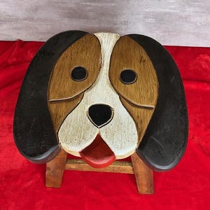 May include: A wooden stool featuring a dog face design on the seat. The dog has black ears, brown patches around the eyes, a white muzzle, a black nose, and a red tongue. The stool is supported by four wooden legs, showing a natural wood finish.