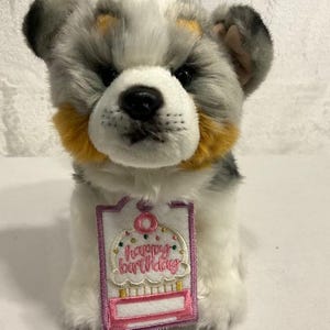 May include: A plush dog toy with gray, white, and yellow fur. It has a black nose and eyes. A tag with the words "happy birthday" and a cupcake design is attached to the toy.