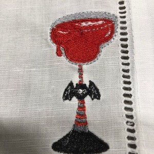 May include: Embroidered white linen cocktail napkin featuring a red cocktail with a gray rim. The stem is black and gray with a bat-like design. The napkin has a decorative cut-out border.