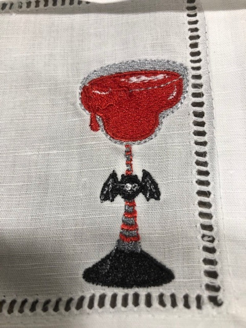 May include: Embroidered white linen cocktail napkin featuring a red cocktail with a gray rim. The stem is black and gray with a bat-like design. The napkin has a decorative cut-out border.