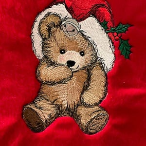 May include: An embroidered brown teddy bear on a bright red fabric background. The bear wears a white and red Santa hat with a small silver bell attached to the brim. A sprig of green holly leaves with red berries is tucked into the hat.