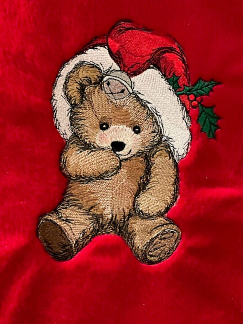 May include: An embroidered brown teddy bear on a bright red fabric background. The bear wears a white and red Santa hat with a small silver bell attached to the brim. A sprig of green holly leaves with red berries is tucked into the hat.