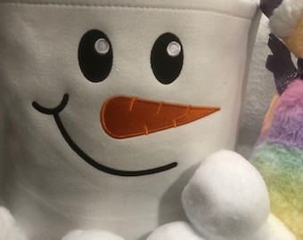 May include: A white snowman-themed bucket with a pink handle, black eyes, an orange carrot nose, and a smiling mouth. Several white, fluffy snowballs are in front of the bucket. A colorful stuffed animal is partially visible on the right.