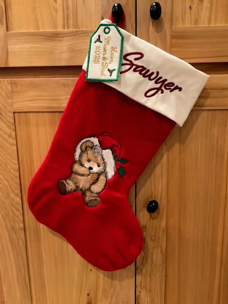May include: A red Christmas stocking with a white cuff embroidered with the name Sawyer in dark red script. The stocking features an embroidered brown teddy bear wearing a red Santa hat and a green holly sprig. A tag reads Love, Mom & Dad 2025.
