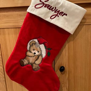 May include: A red Christmas stocking with a white cuff featuring the embroidered name Sawyer in red script. The stocking displays an embroidered brown teddy bear wearing a red Santa hat with green holly leaves and red berries on the red fabric.