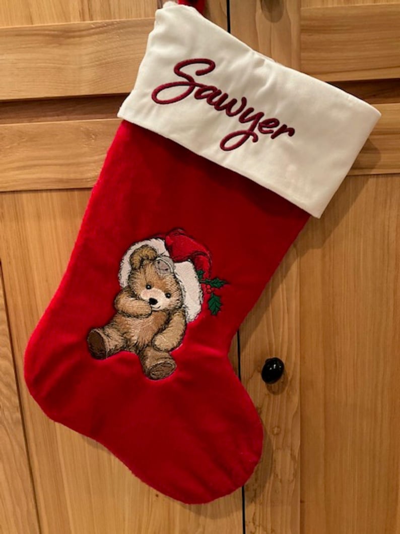 May include: A red Christmas stocking with a white cuff featuring the embroidered name Sawyer in red script. The stocking displays an embroidered brown teddy bear wearing a red Santa hat with green holly leaves and red berries on the red fabric.