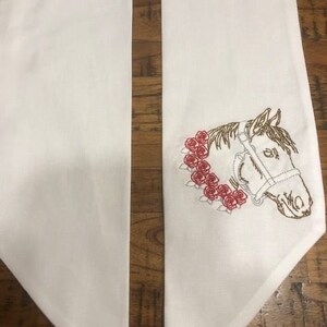 May include: Two white fabric stoles with a gold embroidered horse head design and red roses. The stoles are folded in half and are laying on a wooden surface.