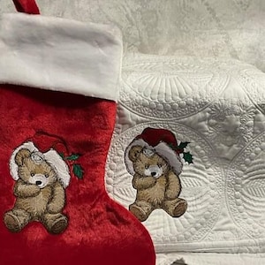 May include: A red velvet Christmas stocking with a white faux fur cuff. The stocking features an embroidered light brown teddy bear wearing a red Santa hat with green holly leaves. A matching embroidered teddy bear patch is placed beside it.