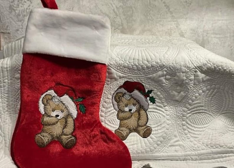 May include: A red velvet Christmas stocking with a white faux fur cuff. The stocking features an embroidered light brown teddy bear wearing a red Santa hat with green holly leaves. A matching embroidered teddy bear patch is placed beside it.