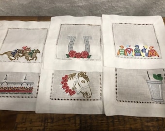 May include: Six white linen napkins with embroidered designs related to horse racing. The designs include a horseshoe with red roses, a jockey riding a horse, a horse's head with red roses, a row of jockeys, a racetrack, and a cocktail glass with a mint garnish.