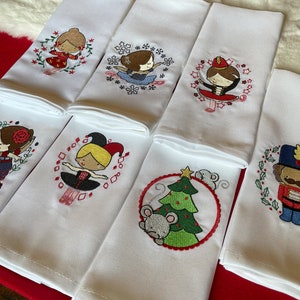 May include: White hand towels with embroidered designs of ballerinas, a Christmas tree with a mouse, and a nutcracker. The ballerina designs feature different outfits and poses, surrounded by floral or geometric patterns. The towels are folded and arranged on a red surface.