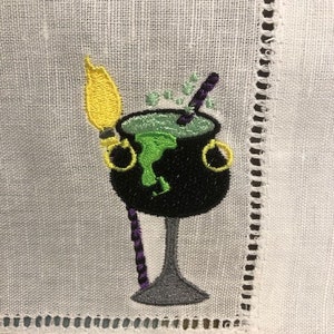 May include: Embroidered white linen napkin featuring a black cauldron with green bubbling liquid, a yellow broom, and a purple stirring stick. The cauldron has gold handles. The napkin has a decorative stitched border.