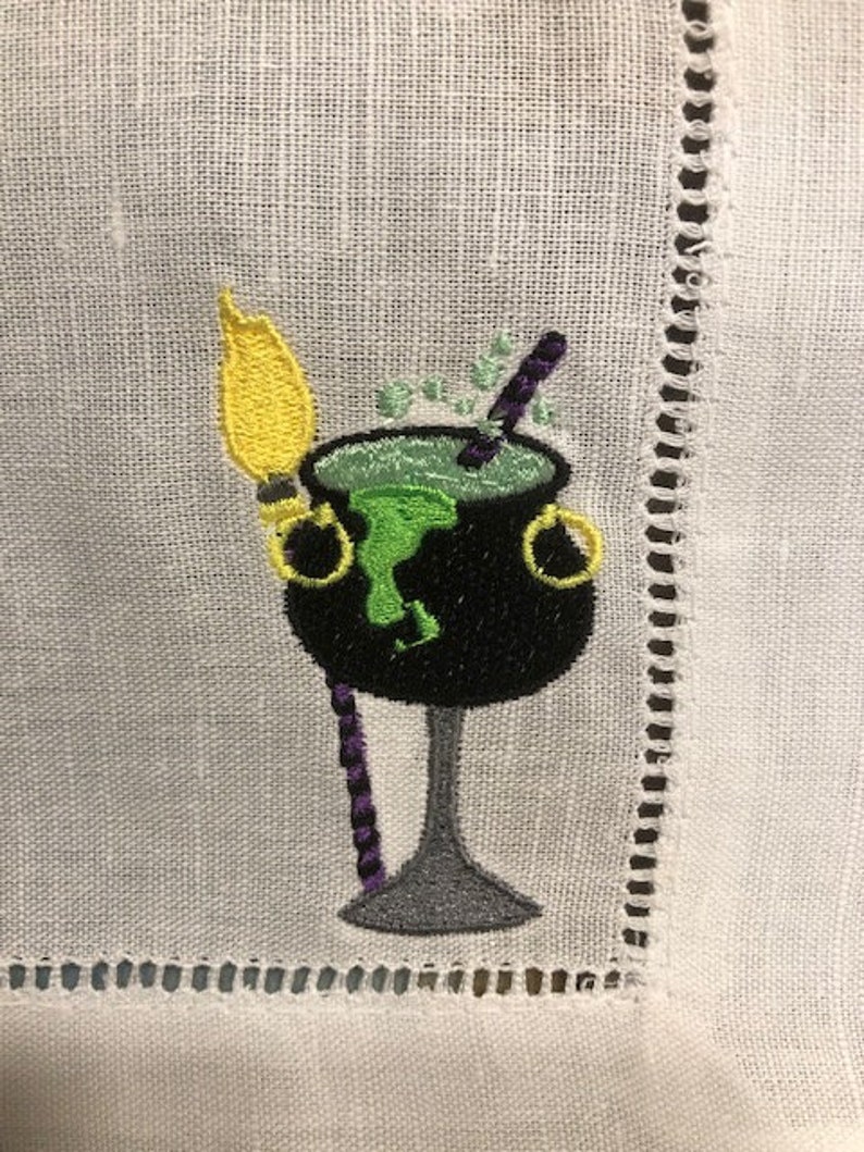 May include: Embroidered white linen napkin featuring a black cauldron with green bubbling liquid, a yellow broom, and a purple stirring stick. The cauldron has gold handles. The napkin has a decorative stitched border.