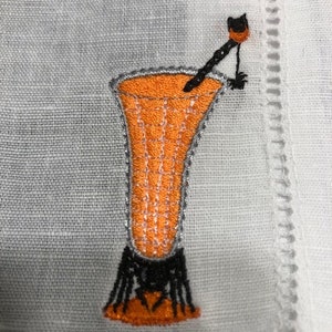 May include: Embroidered cocktail glass design in orange, black, and silver on a white linen napkin. The cocktail glass has a detailed design with a black stem and a decorative stirrer. The napkin has a decorative border.