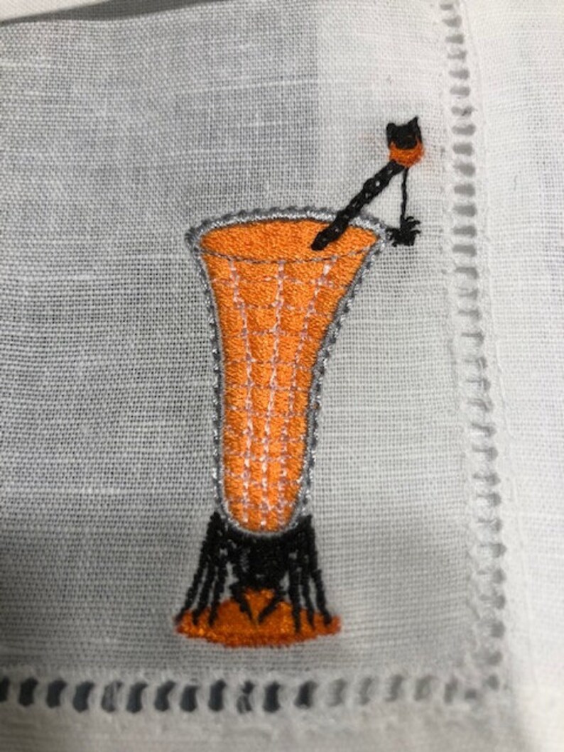 May include: Embroidered cocktail glass design in orange, black, and silver on a white linen napkin. The cocktail glass has a detailed design with a black stem and a decorative stirrer. The napkin has a decorative border.