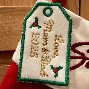May include: A white felt gift tag with a green stitched border and a green eyelet. The tag features gold embroidered text reading Love Mom & Dad 2026, accented by two small green holly leaf motifs with red berries, attached to a red and white stocking.