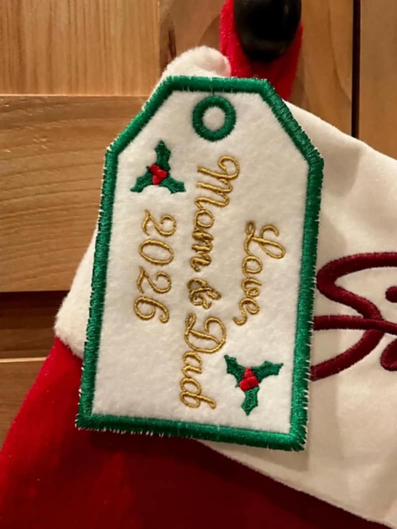 May include: A white felt gift tag with a green stitched border and a green eyelet. The tag features gold embroidered text reading Love Mom & Dad 2026, accented by two small green holly leaf motifs with red berries, attached to a red and white stocking.