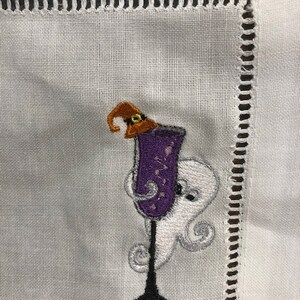 May include: White linen cocktail napkin with an embroidered Halloween design. The design features a purple cocktail glass with a witch's hat, and a friendly ghost. The napkin has a decorative stitched border.